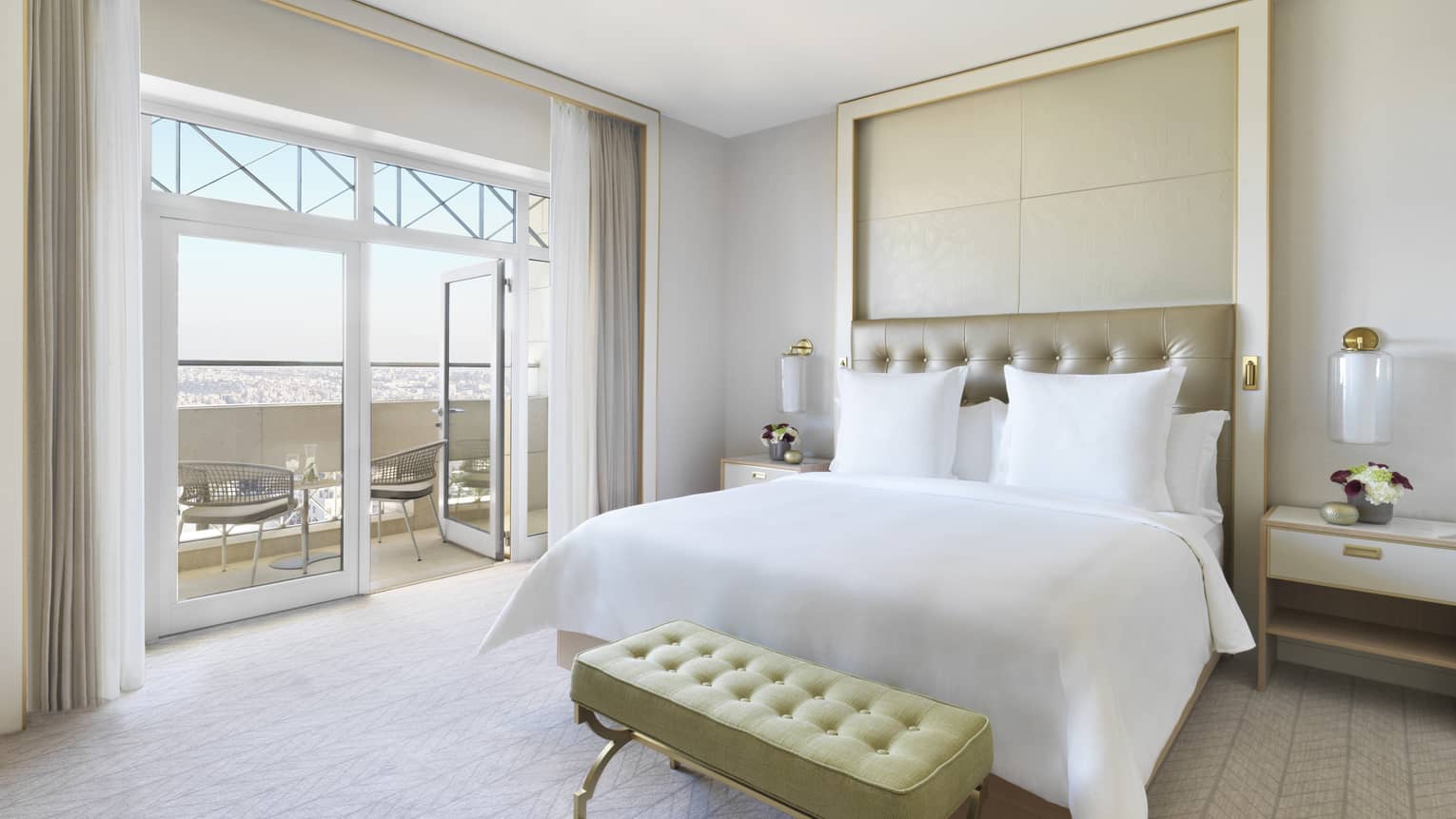 A plush white bed in the centre of the room with a yellow ottoman at the foot. The room opens up to a veranda overlooking the city. 