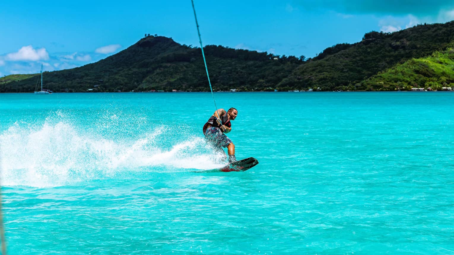 A guest creates a spray of water with their wakeboard as they're towed through the ocean before a backdrop of forested hills.