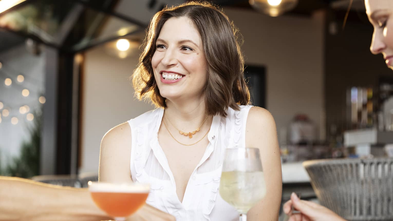 Person with brown shoulder-length hair wears a white sleeveless button-down shirt and sits smiling at a table filled with drinks and food and two other people on either side