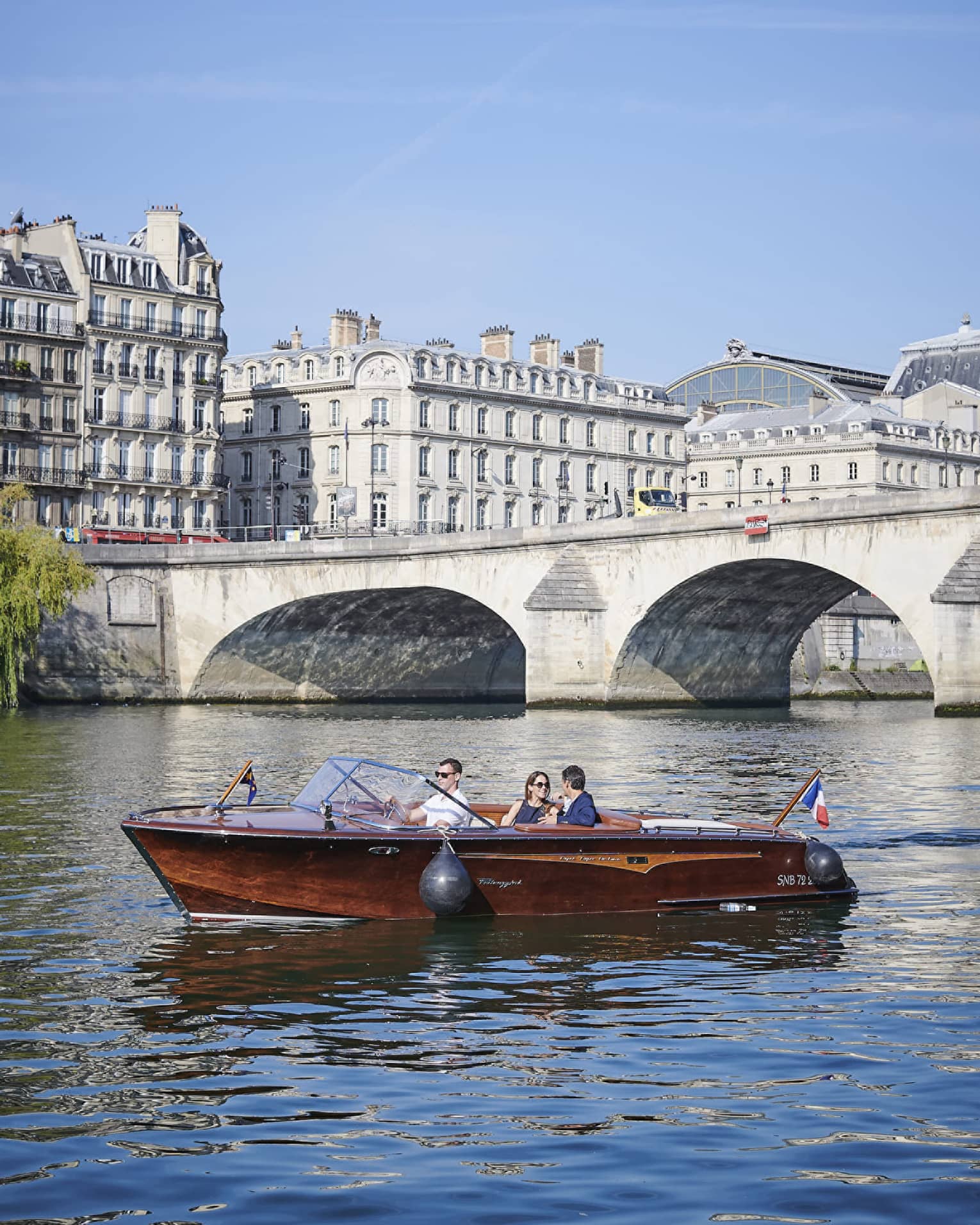 A couple relax in a brown speedboat cruising near a stone bridge along a river lined with white Haussmannian-style buildings.