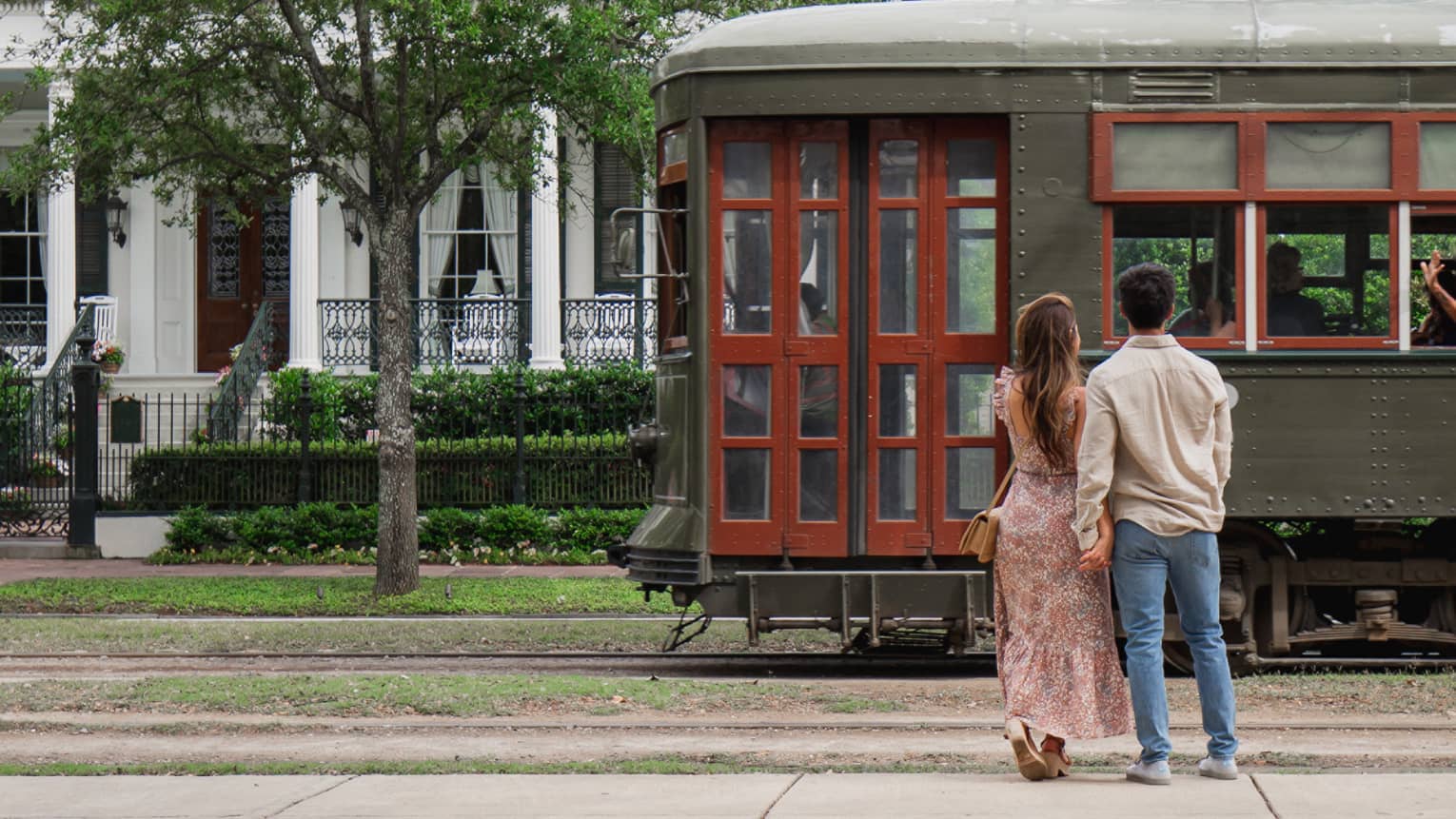 Woman and man hold hands in front of old-fashioned streetcar on New Orleans street, white house in background