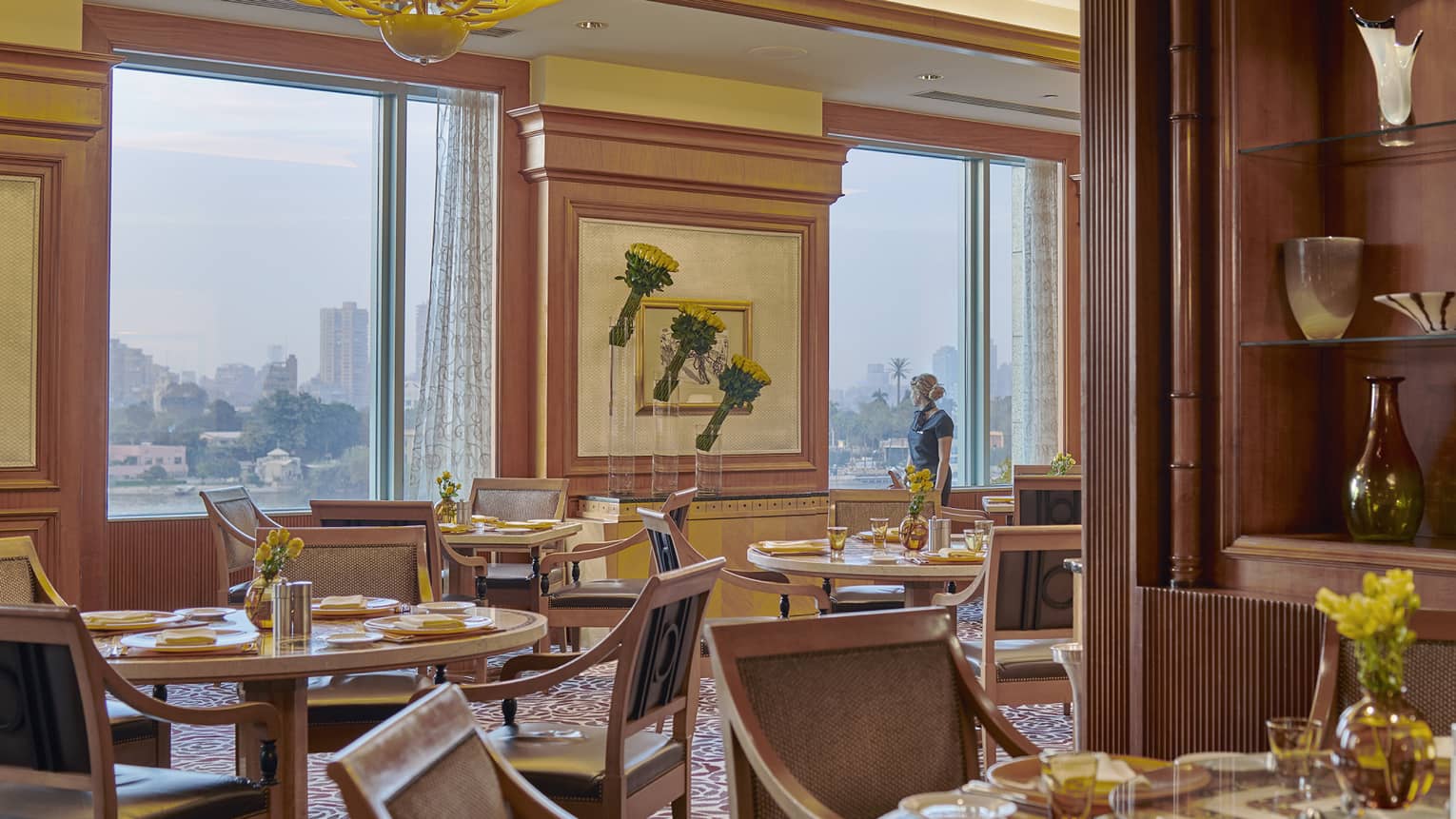Hotel staff stands at window overlooking river and city in dining room beyond tables, chairs 