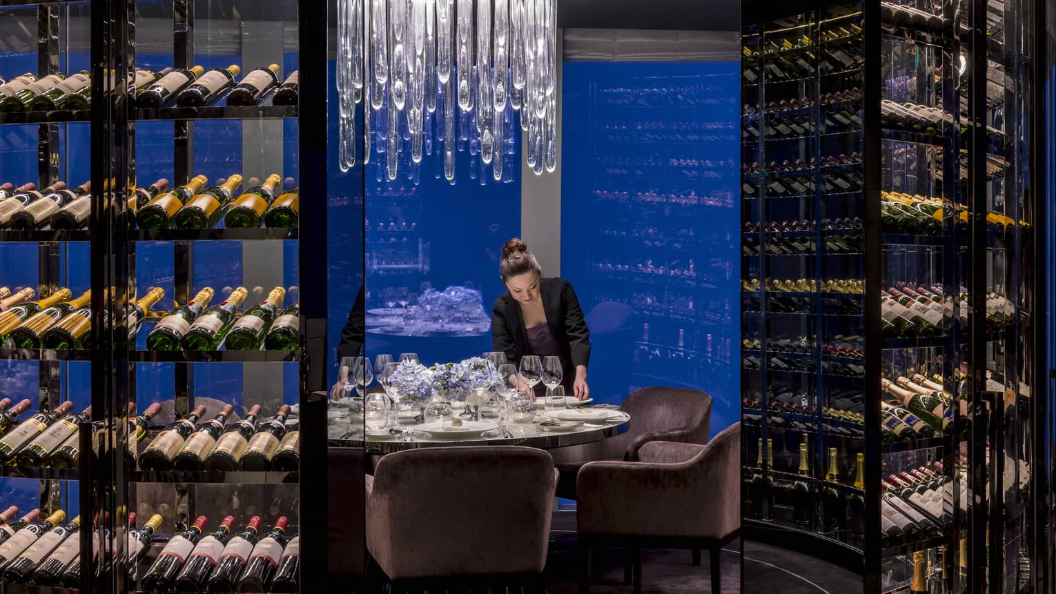 Woman sets plate on round private dining table under crystal chandelier, surrounded by wine cellars