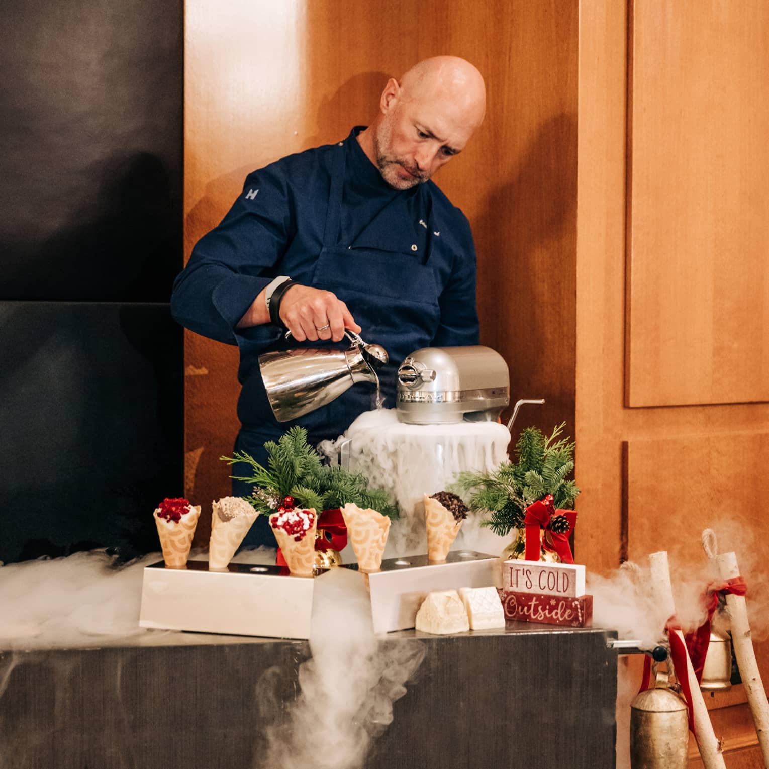 Chef pours liquid nitrogen into a mixer in front of a cart topped with waffle cones and holiday decor