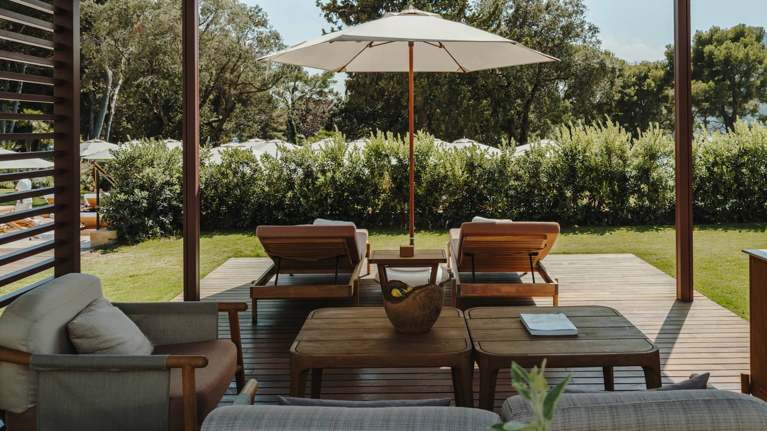 A shaded pool cabana with comfortable seating, a table, lounge chairs, an umbrella and lush green surroundings.