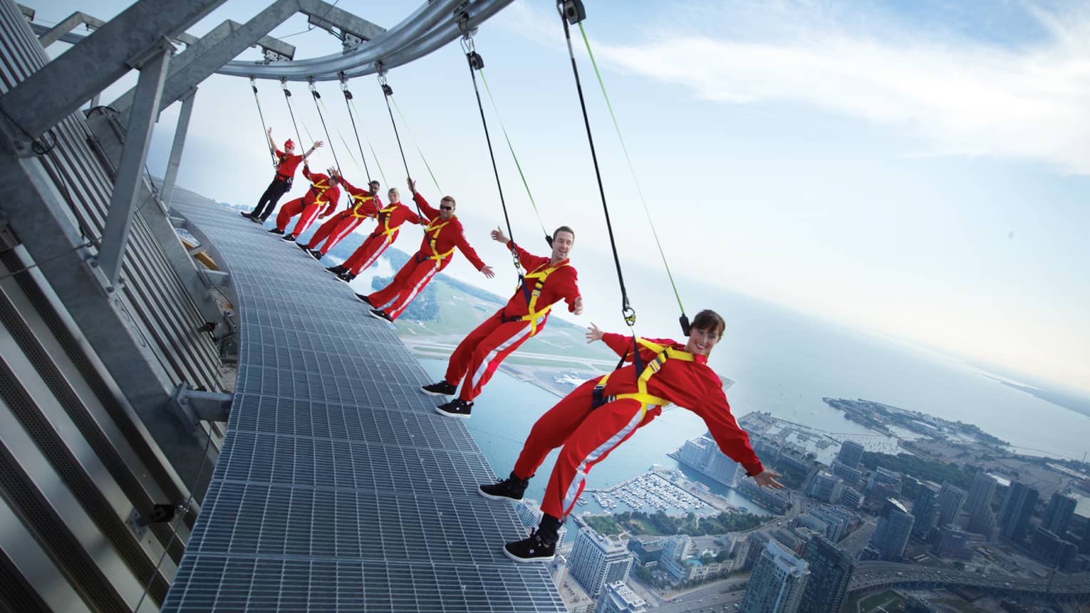 People wearing red jumpsuits, harnesses lean back over edge of CN Tower high above city