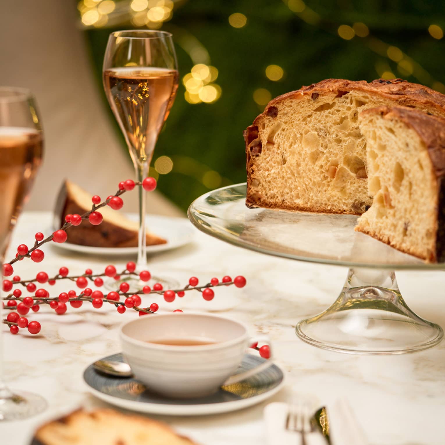 Loaf of panetonne sits on a crystal cake stand with two slices plated on the table next to it, two glasses of pink-hued wine in champagne flutes and a cup of tea