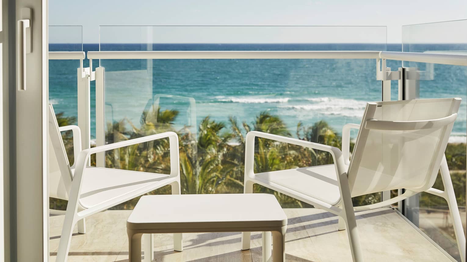 Luxury hotel balcony with two white chairs and a table overlooking the ocean and palm trees under a clear blue sky