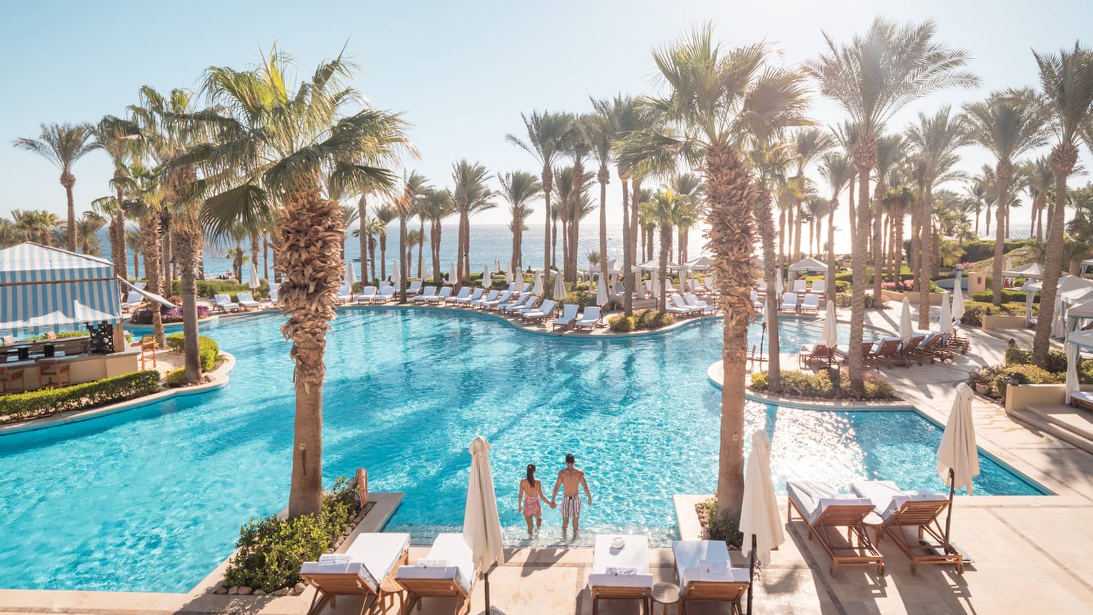 Couple enters outdoor swimming pool under palm trees, sun