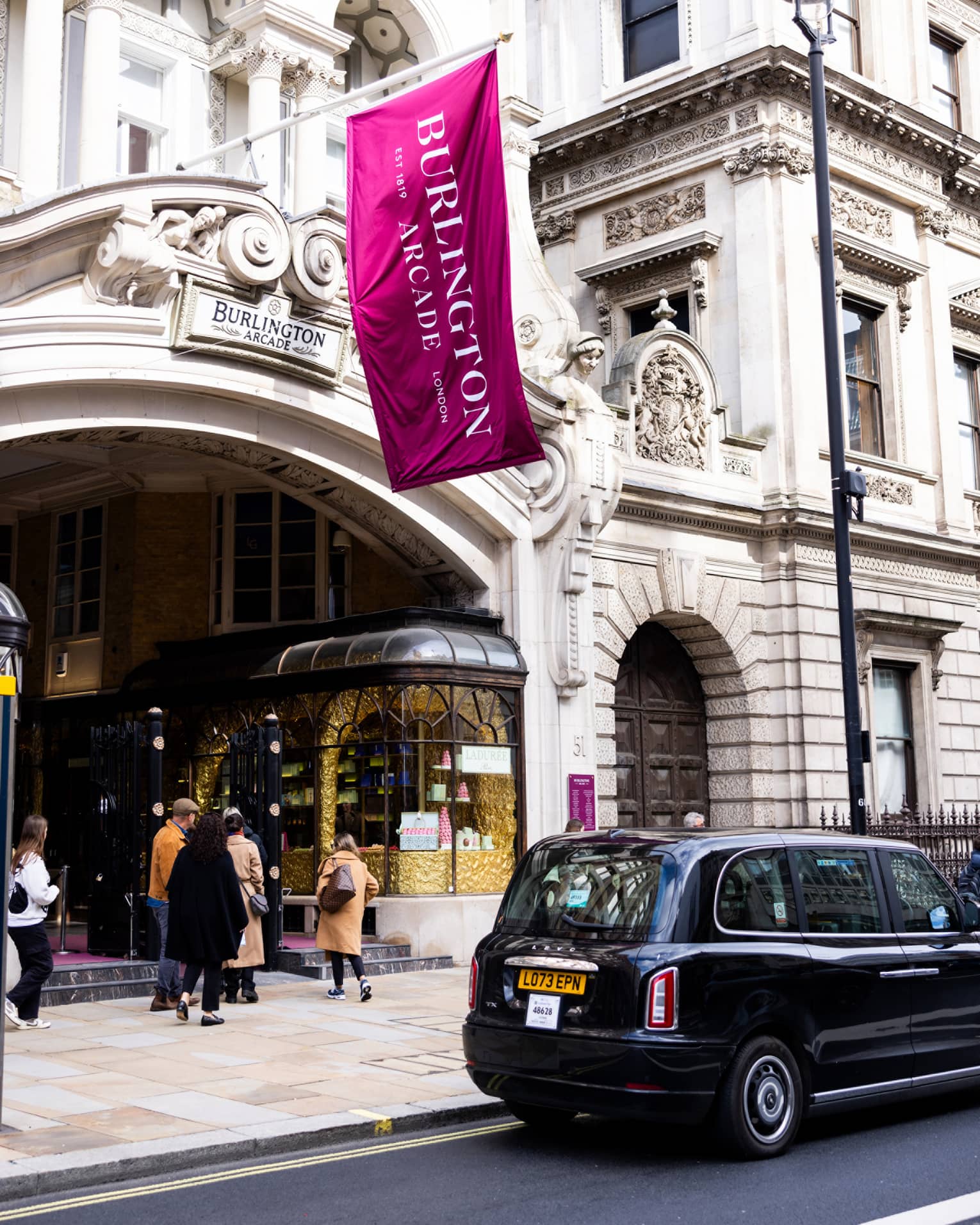 London-style black cab parked in front of a high-end pastry shop in a building with a Burlington Arcade sign and flag.