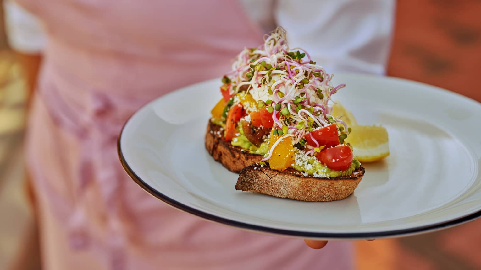 Chef wearing a pink apron and holding a plate with two slices of bread with greens and vegetables on it and a lemon slice on the side.