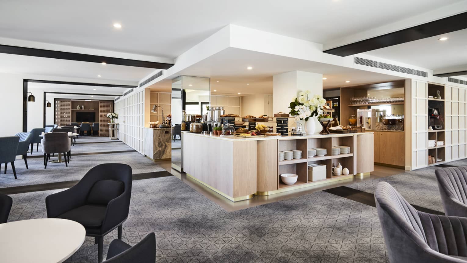 A modern hotel lounge with plush grey chairs and carpet, and a well-equipped kitchen set up with buffet foods and coffees.