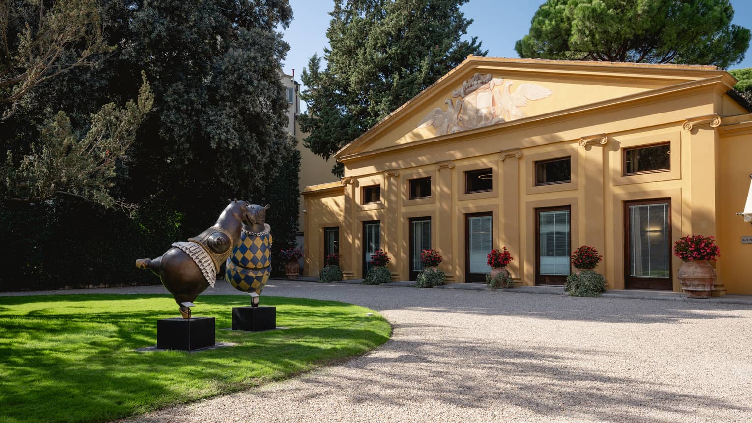 A small building with large windows on manicured grounds complete with potted plants, a drive and sculptures, with large trees in the background