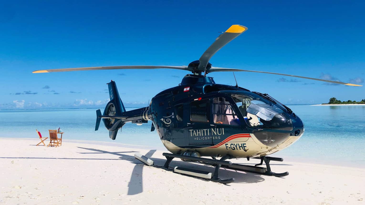 A helicopter parked on the beach beside a calm ocean; nearby a small table and chairs are turned towards the horizon.