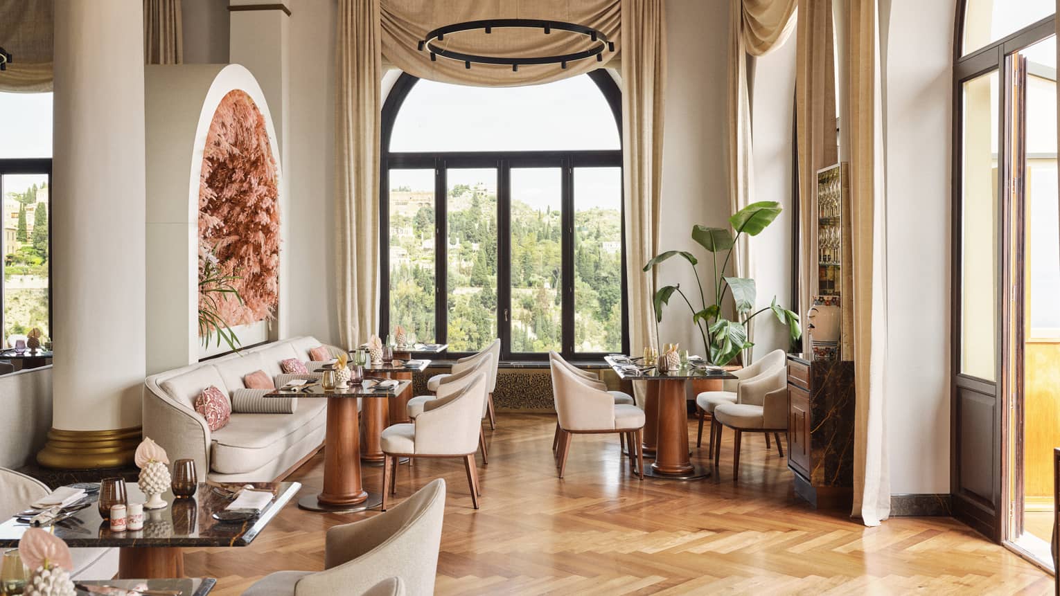 Restaurant dining room with parquet wood flooring, tall ceilings, white columns, banquet seating and large windows letting in natural light