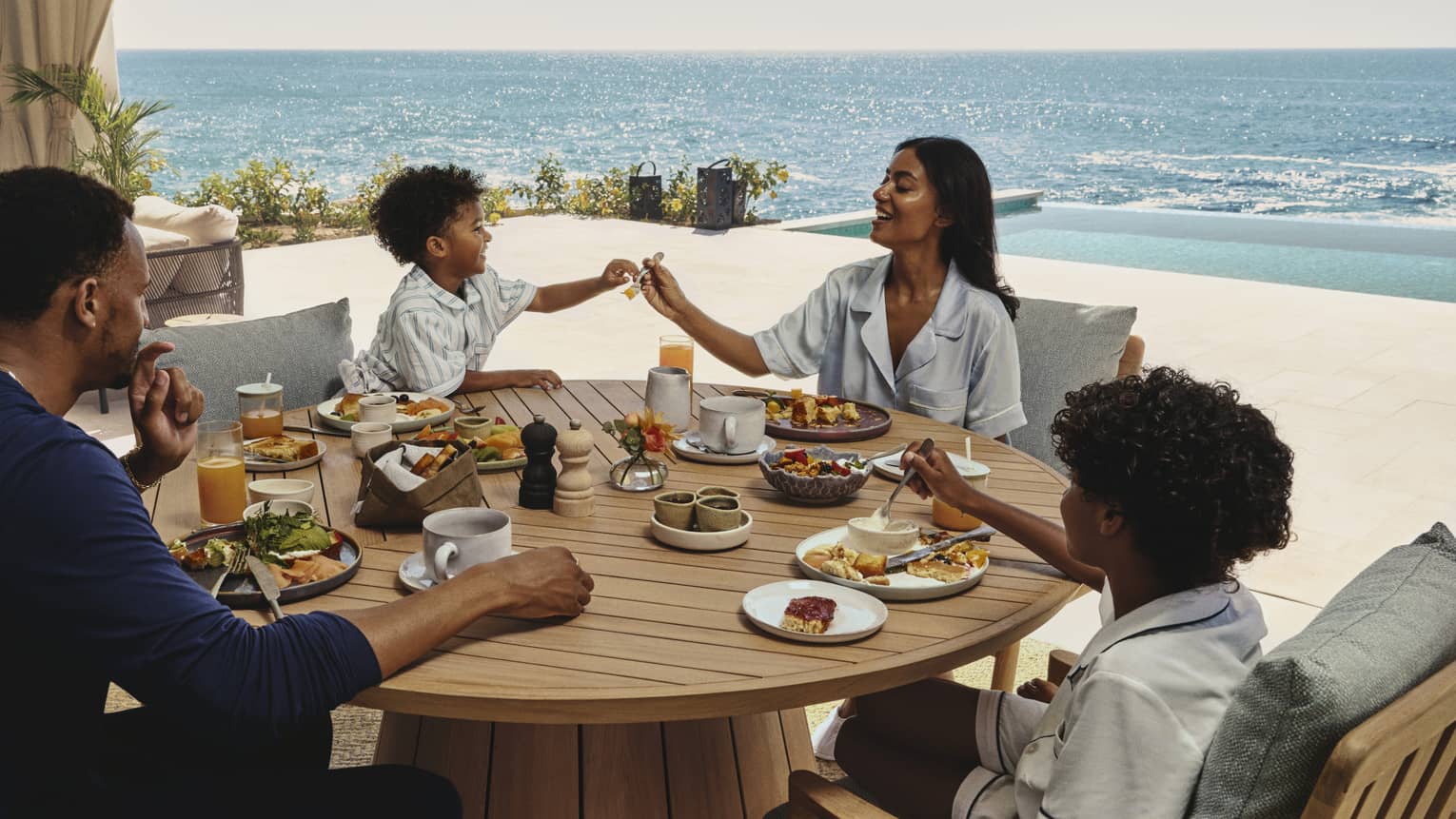Family enjoying breakfast together at a round wooden table on an ocean-view terrace at a luxury resort
