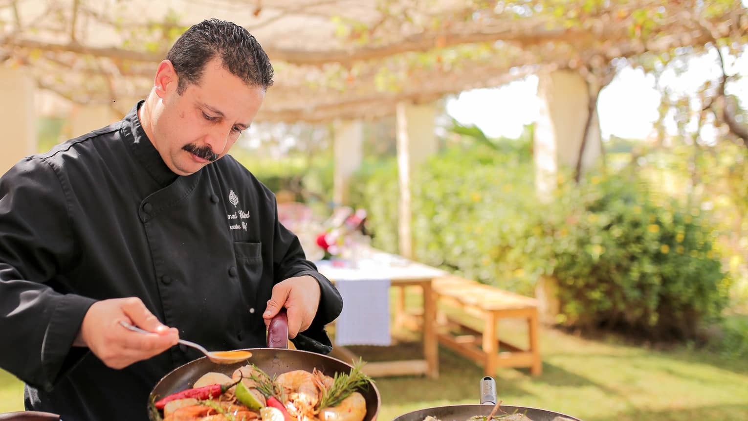 Chef Emad Ebeid pours sauce over seafood dish in pan at outdoor kitchen