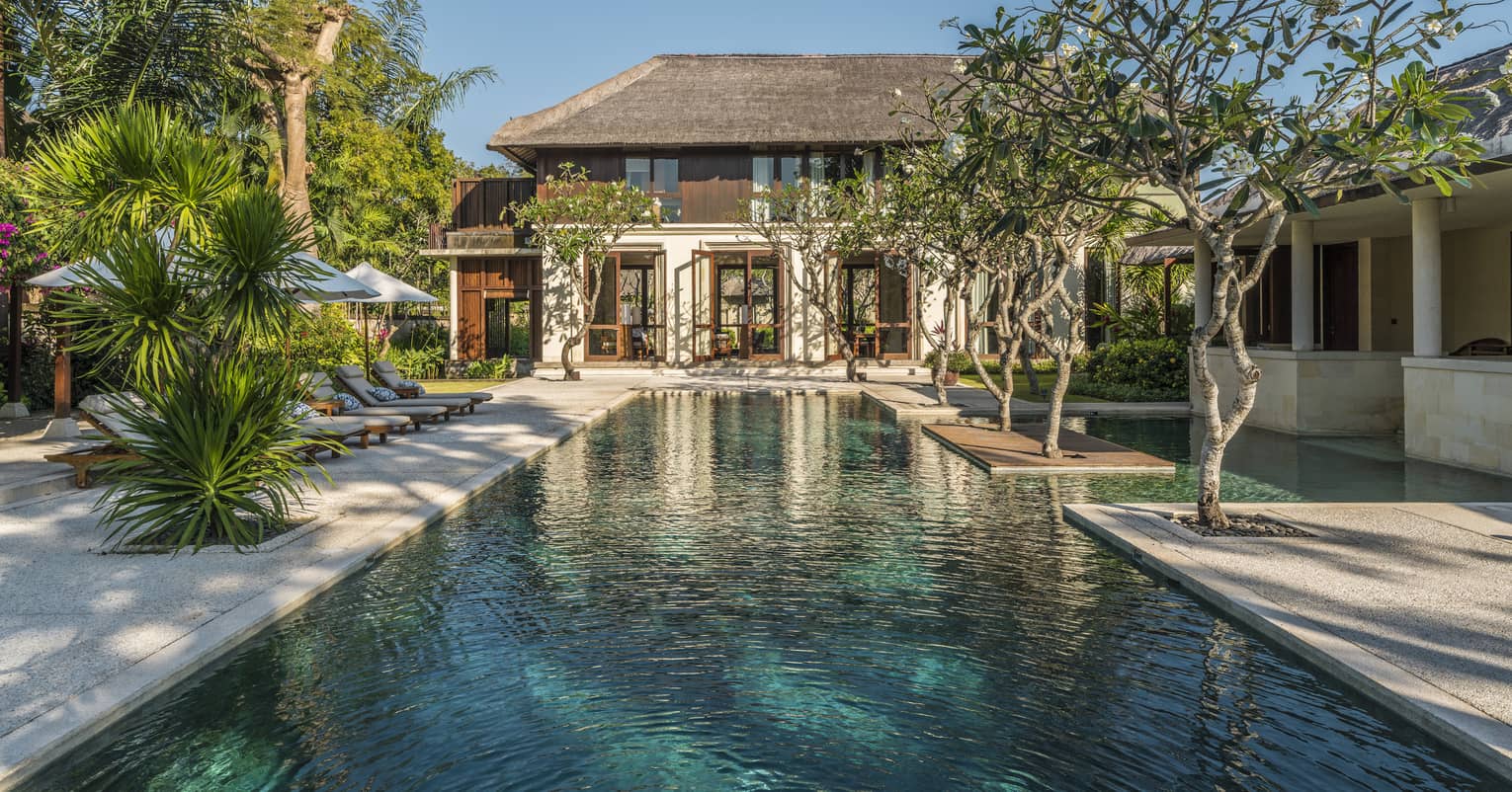 Two-storey villa at end of long outdoor swimming pool lined with trees, tropical plants