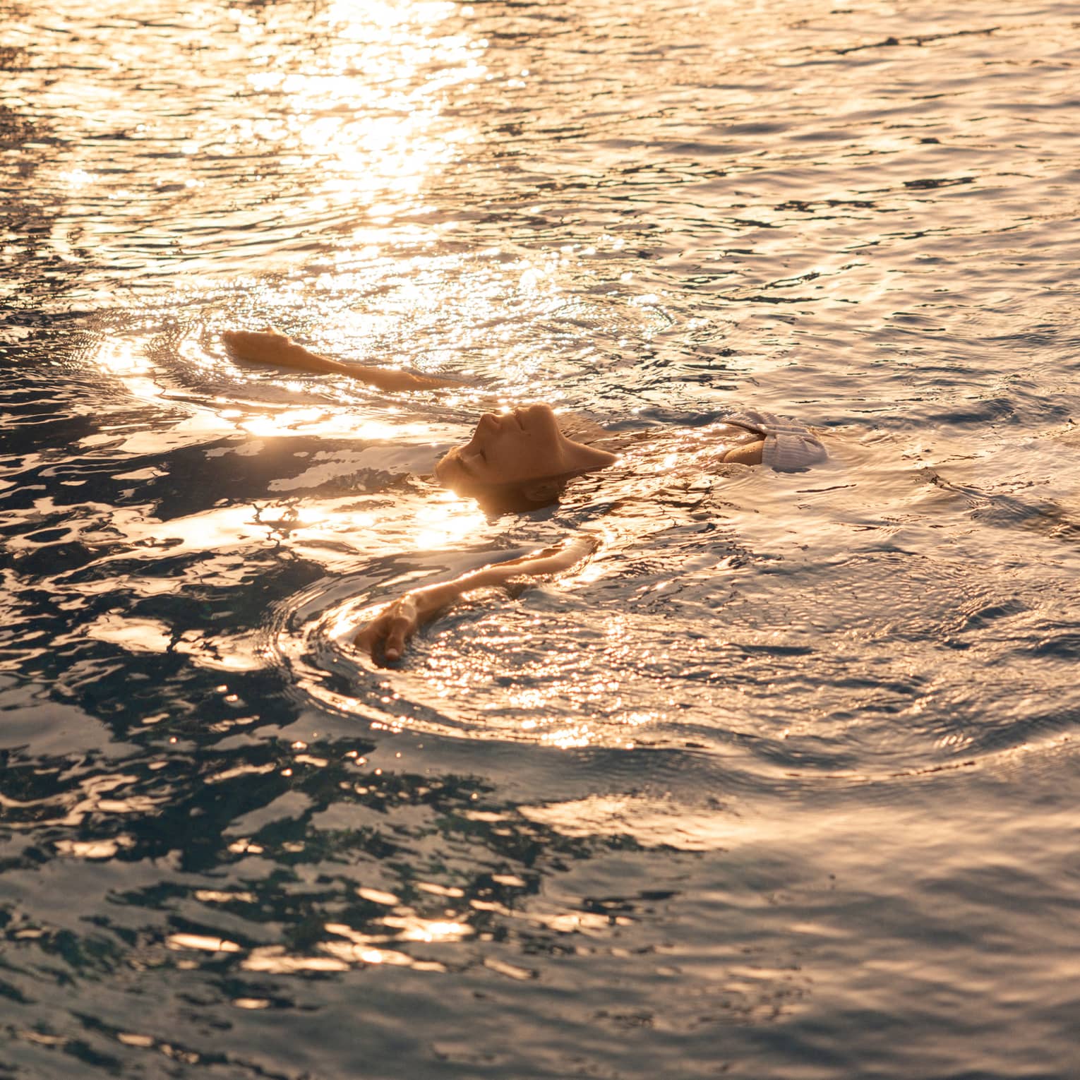 A person floating on their back in the ocean, with sunlight rippling across the water.