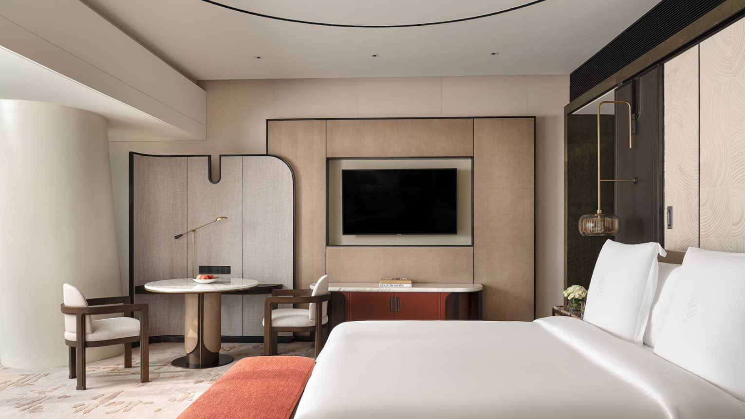 Contemporary hotel room with king bed, red cushioned bench, round table with two chairs and TV