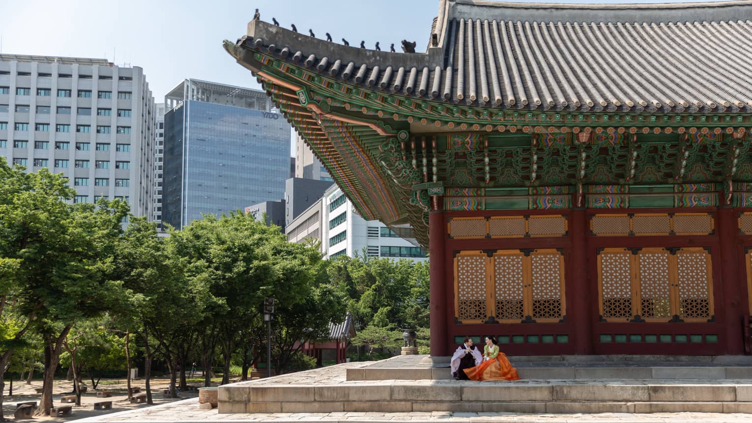 A couple in traditional Korean dress are seated on stone steps under the colourful roof awning of a tree-lined palace.
