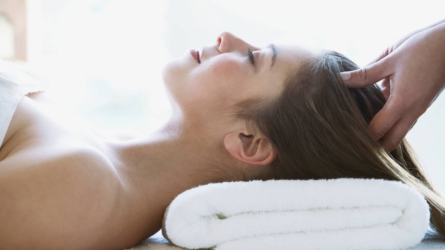 Woman lays on back with head on folded white towel, closes eyes as hands massage her scalp