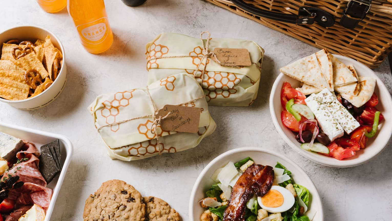 An assortment of picnic food including salad, cookies, sandwiches, fruit, and sliced meat next to a brown wood basket.