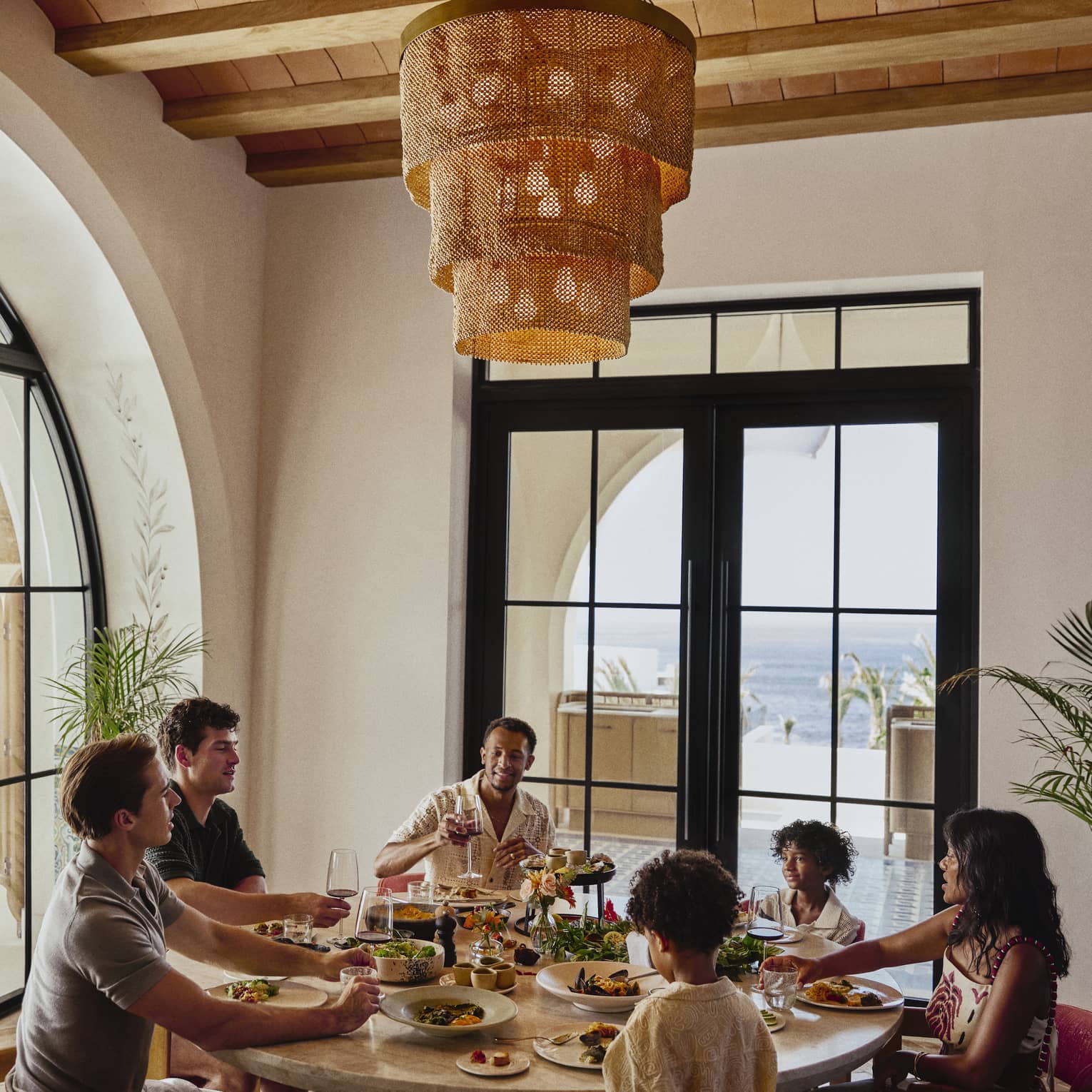 A six-person family enjoys a colourful meal in an airy room. Behind them, French doors lead to a patio overlooking the ocean.