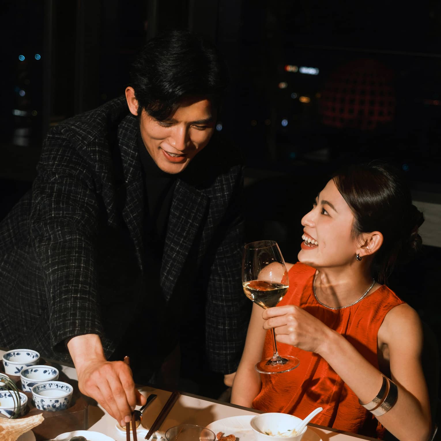 Guest reaches into a hot pot with chopsticks while another smiles holding a glass of white wine. Behind them is a night sky.