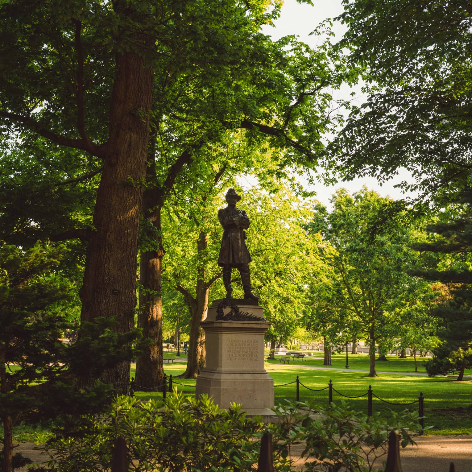 A bronze statue of Thomas Cass, called Colonel Thomas, shaded by greenery from the bright sun in Boston’s Public Garden.