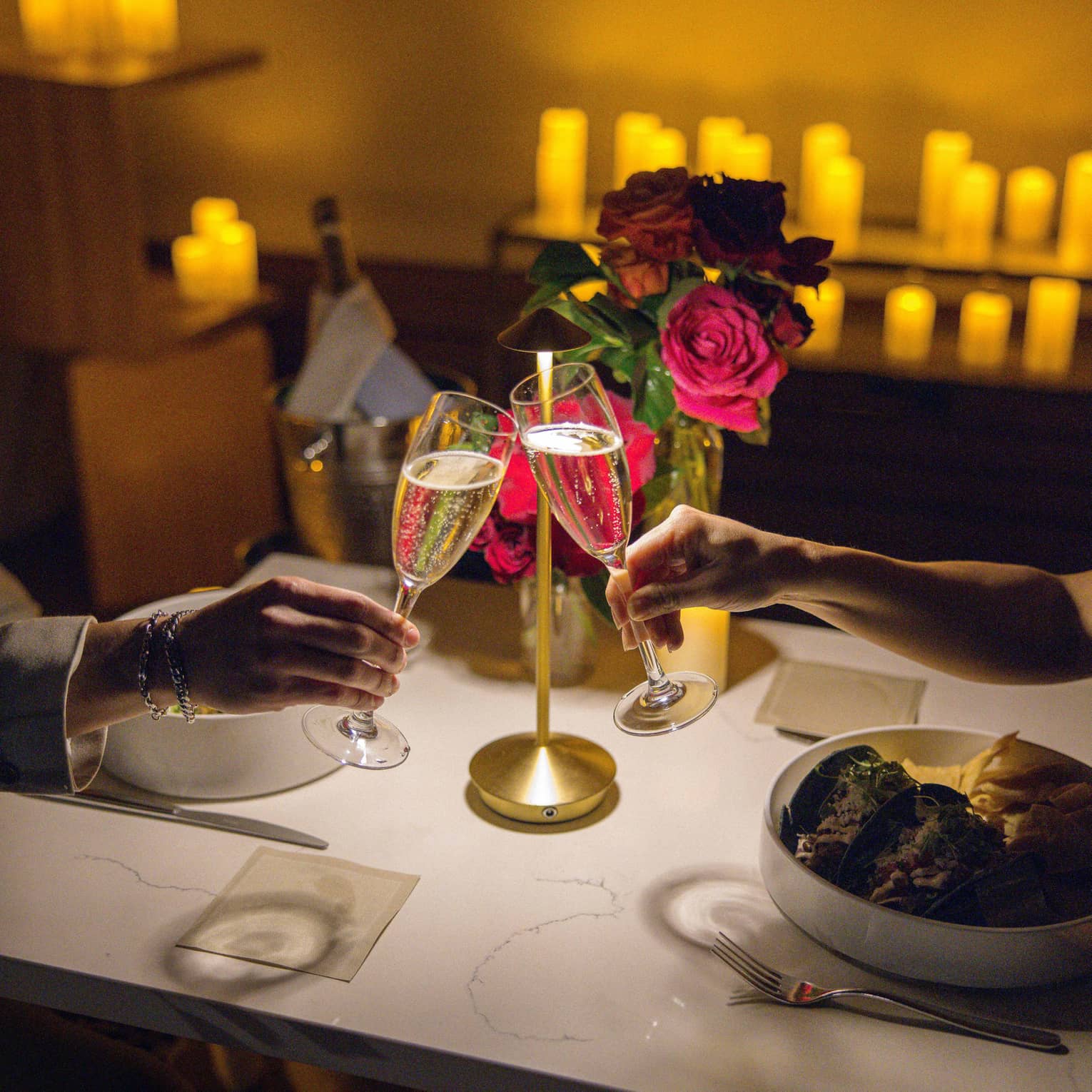 Two hands clink champagne glasses over a dining table set with flowers, candles in the background