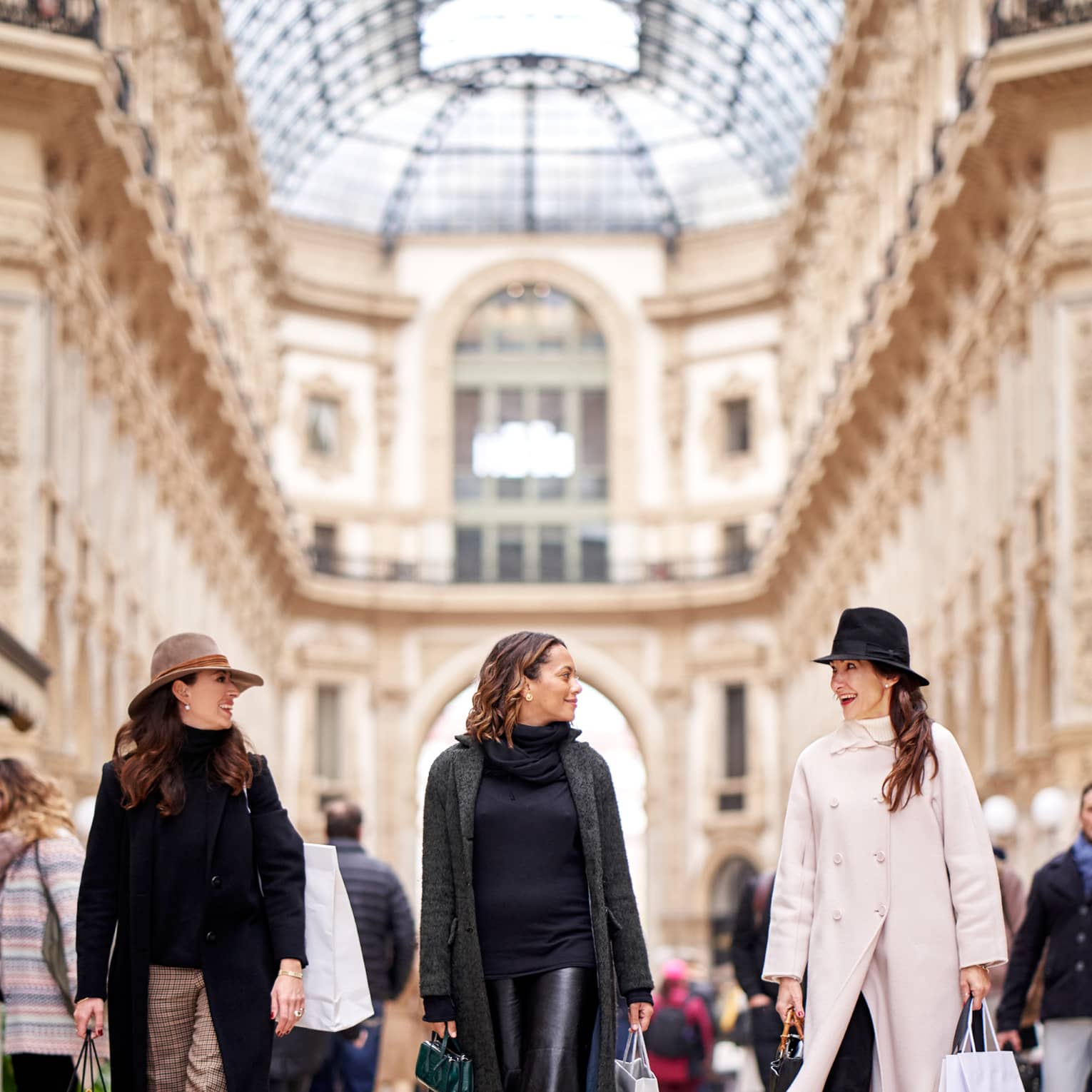 Front view of three chic shoppers smiling at each other as they stroll through the airy arcade of the shopping galleria.