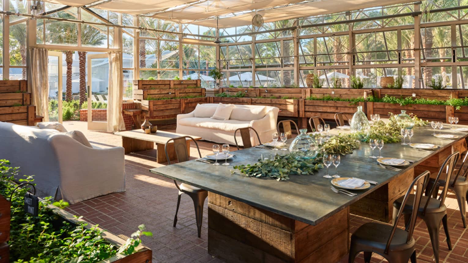 Glass-walled gathering space with a long rectangular table with metal bistro chairs, place setting for 8 and a green garland centrepiece on one end of the room and two white sofas and a coffe table on the other side of the room