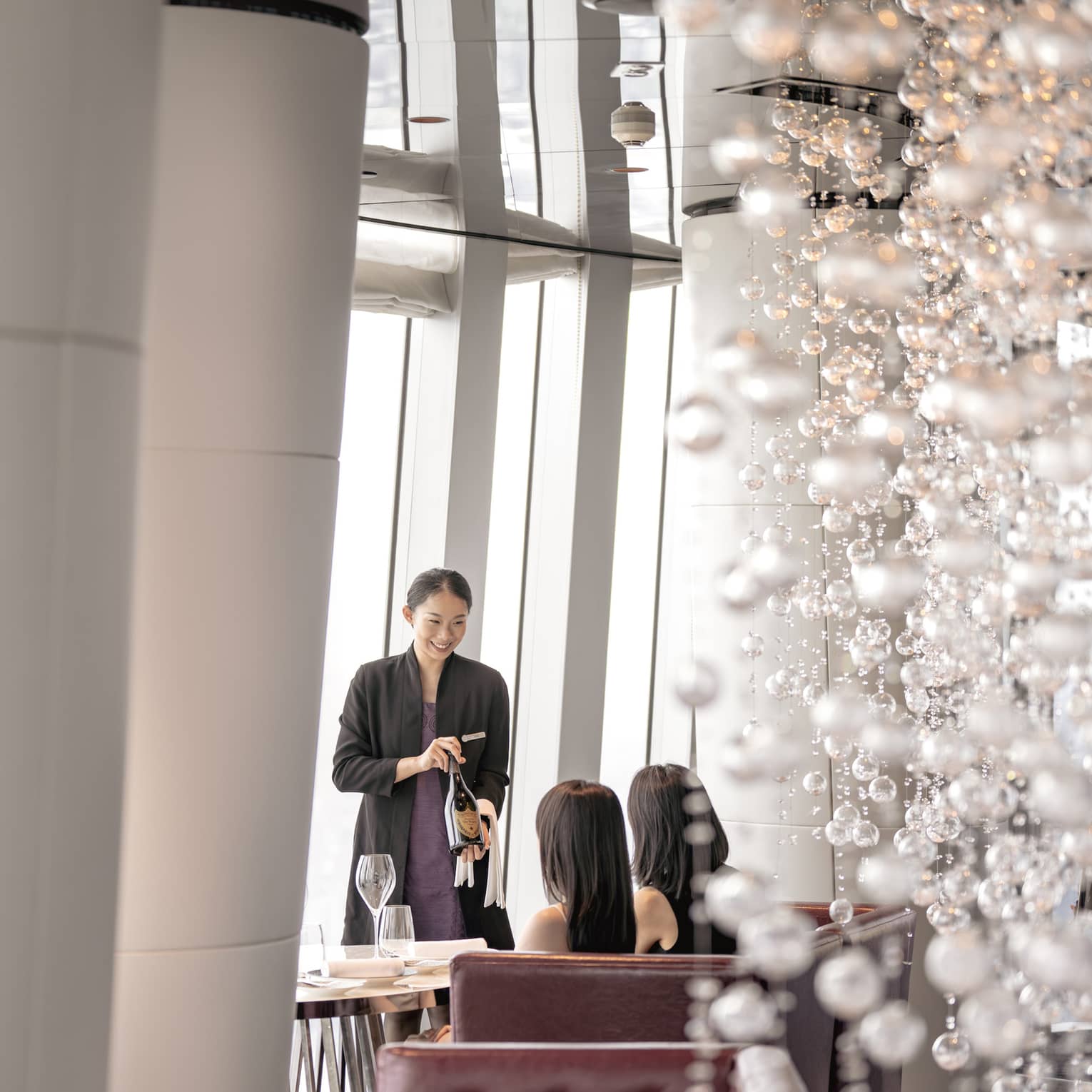 A server presents a bottle of wine to two diners in a leather tufted banquette, a cascade of shimmering white baubles behind.