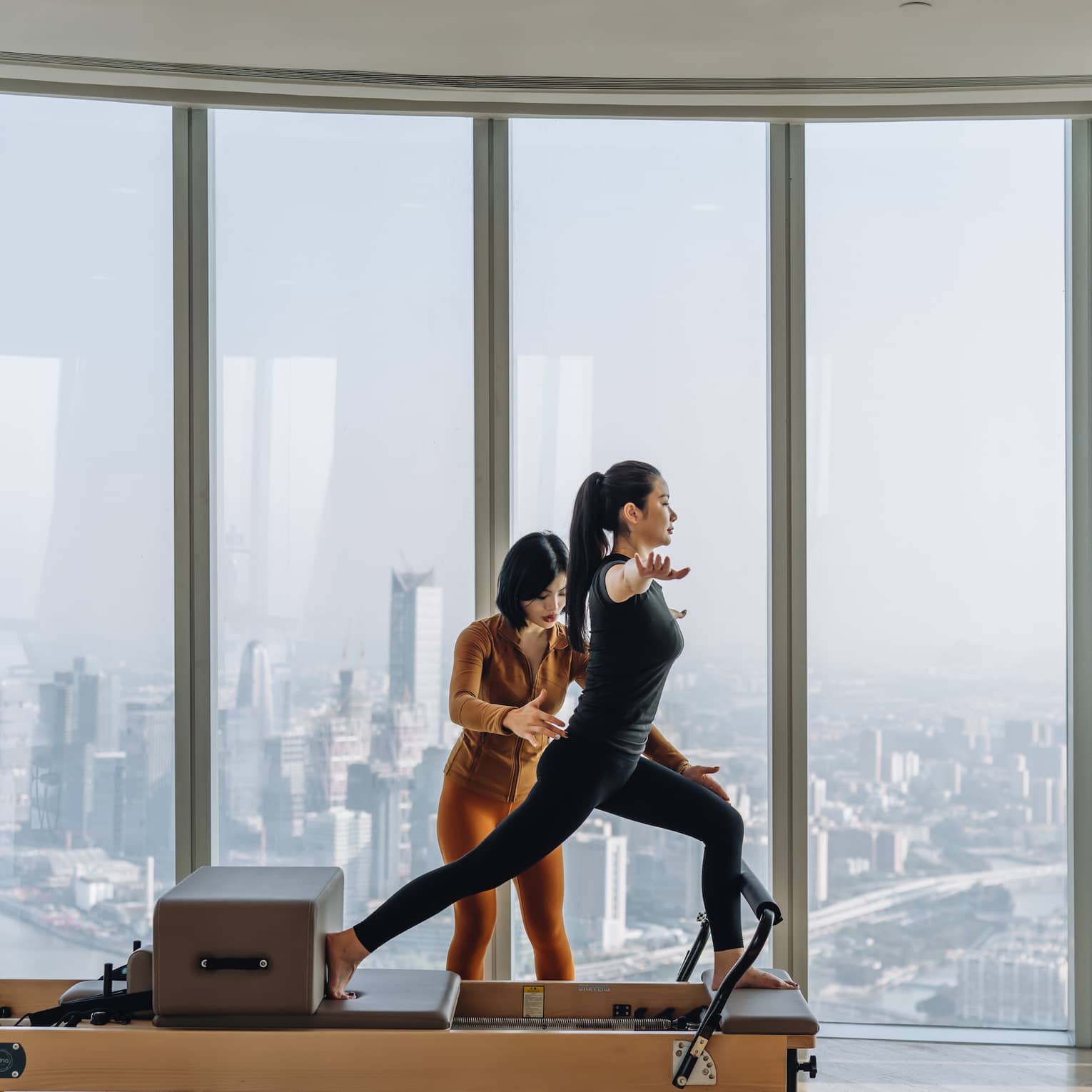 An instructor gently adjusts the positon of a guest in a lunge on a reformer machine, high up overlooking a hazy cityscape.