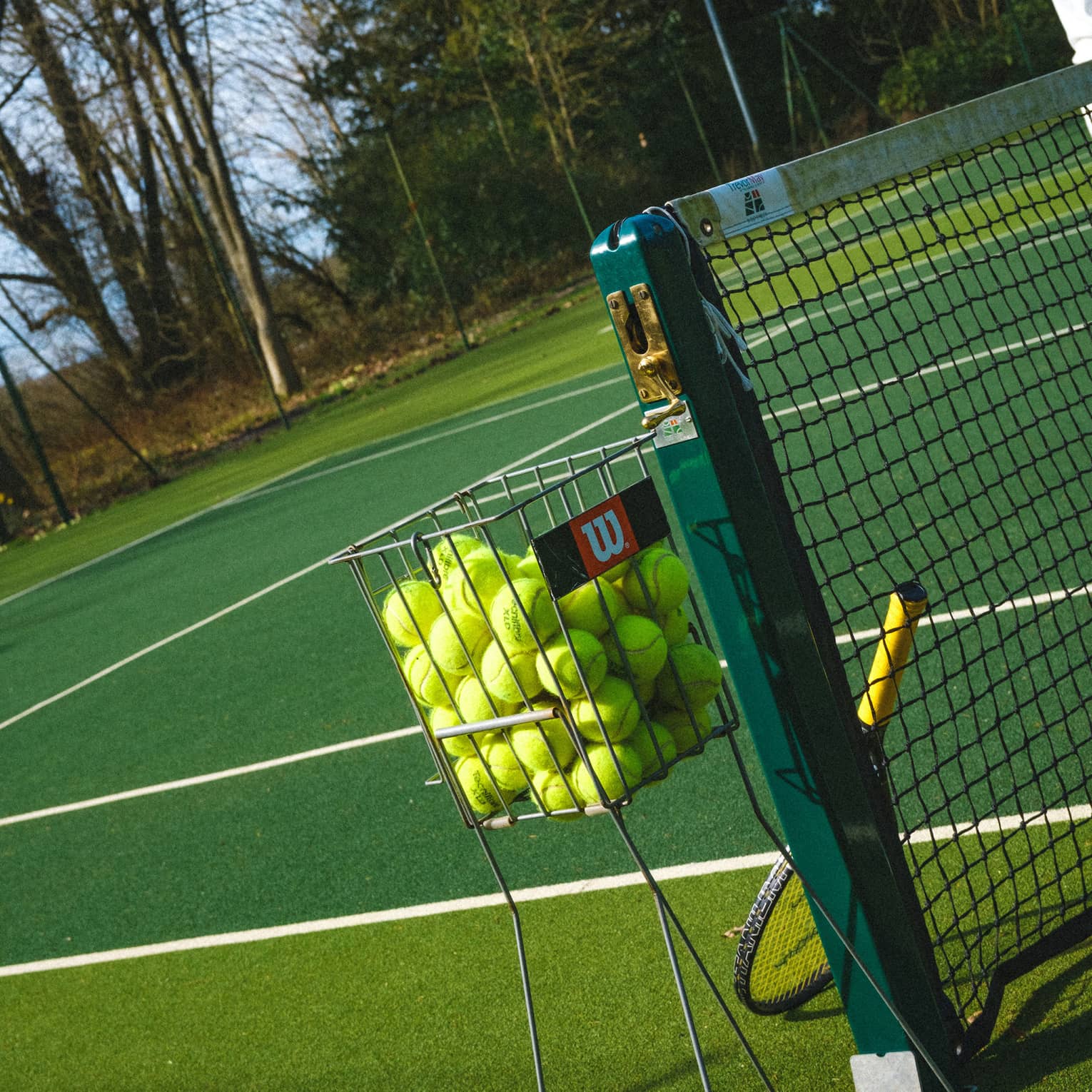 Tilted close-up of a basket filled with yellow tennis balls beside a net upon which leans a racquet on a green court.
