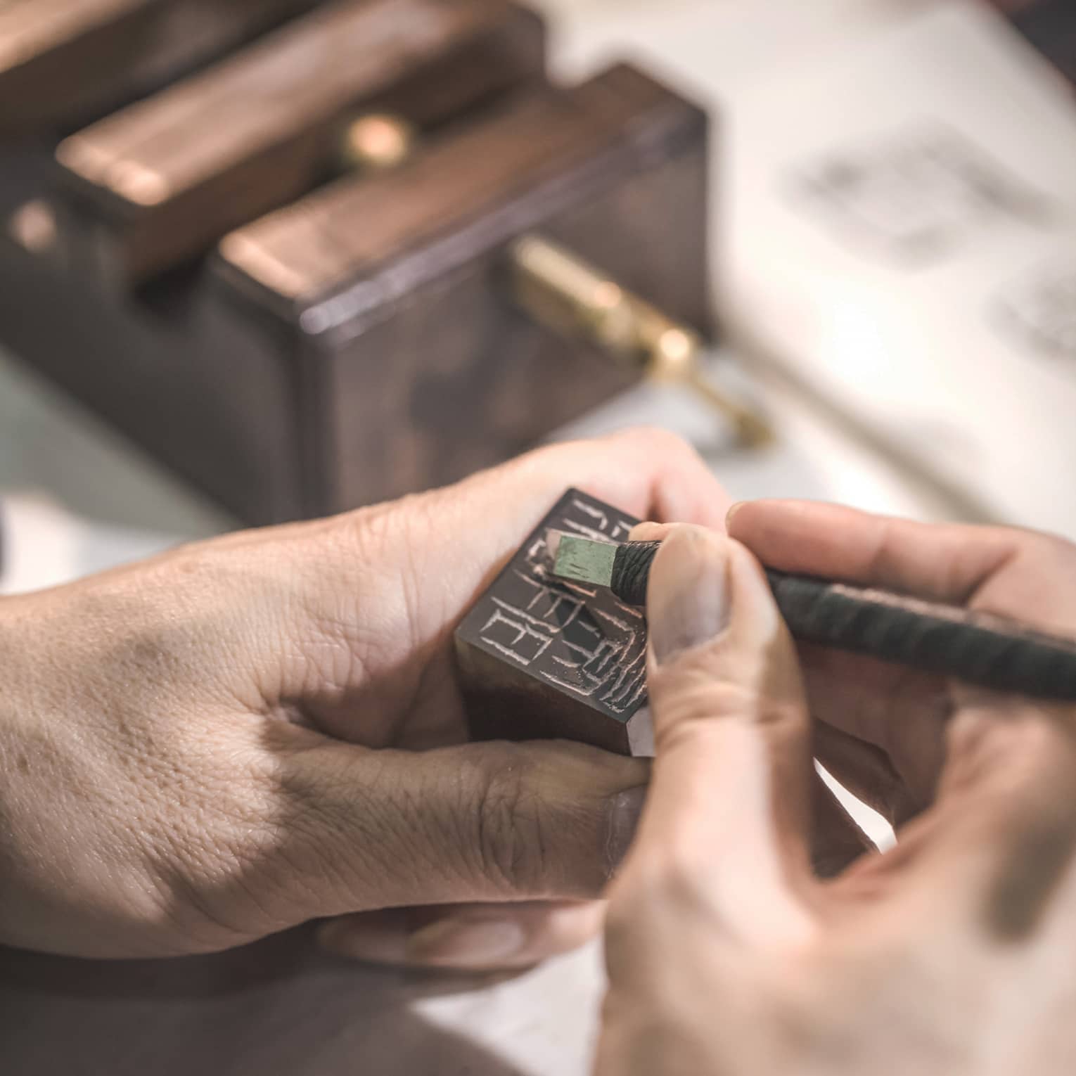 A hand holds a small block of dark stone while another hand carefully uses a tiny chisel to carve symbols into its surface.