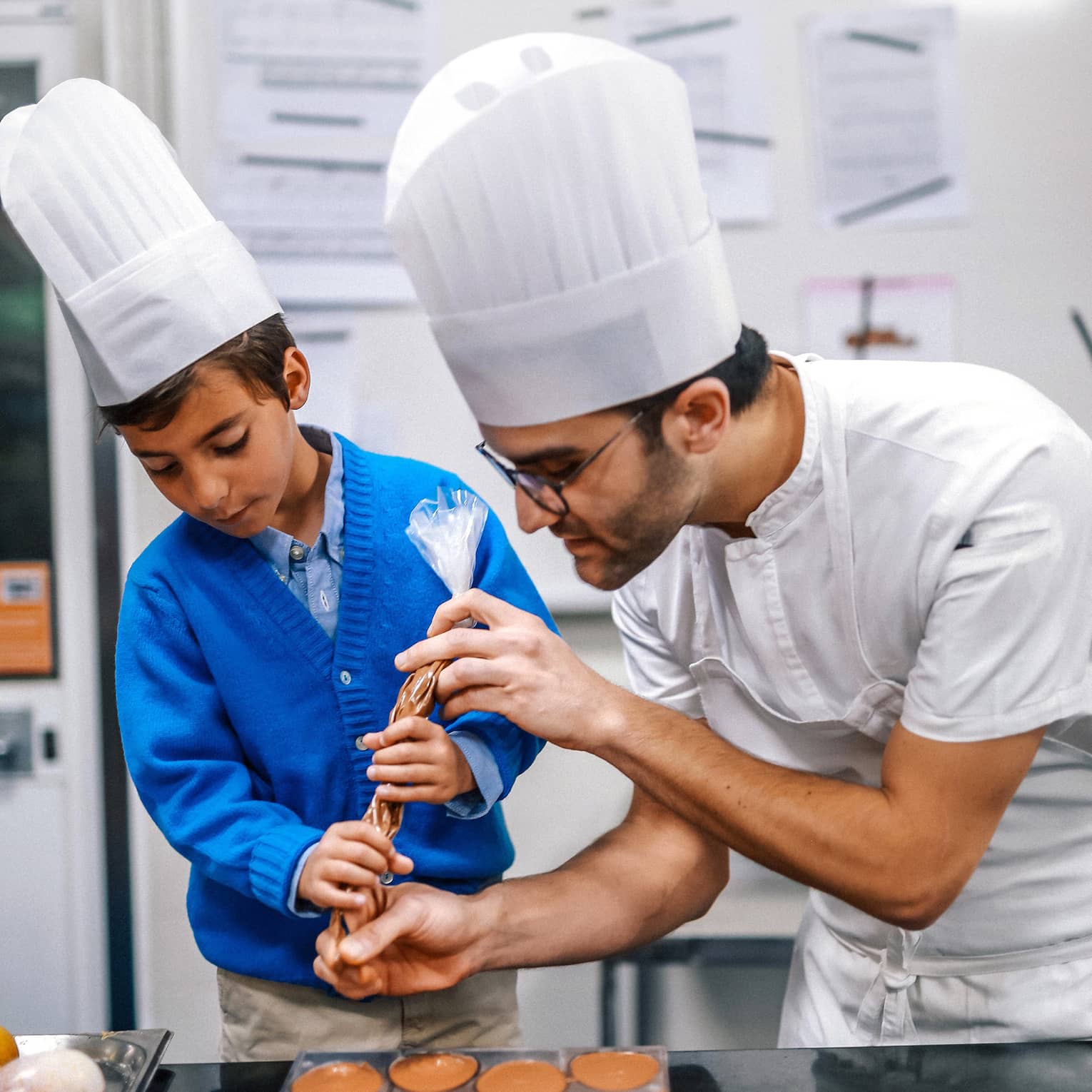 A pastry chef and a child, both in chef's hats, squeeze chocolate circles from a plastic piping bag onto a small platform.