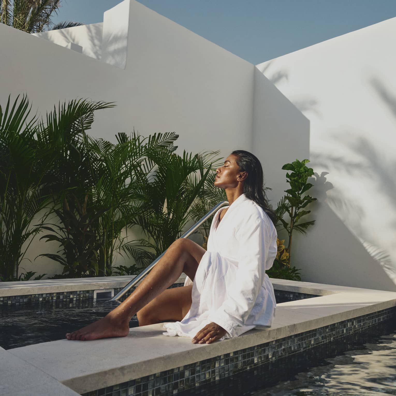 A guest in a linen bathrobe soaks in the sunlight while dipping their feet in a tiled pool surrounded by walls and plants.