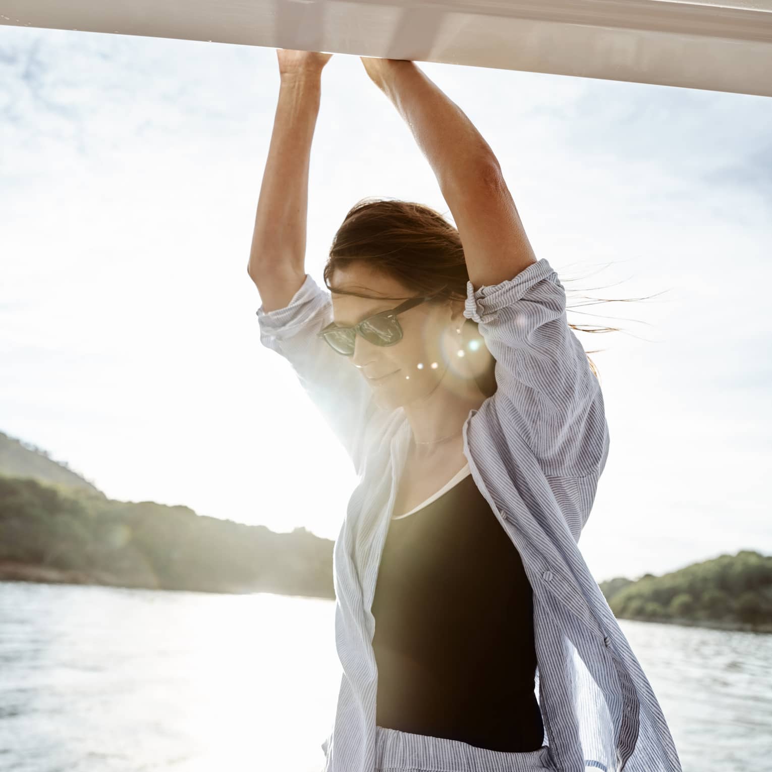 Close-up of a guest with arms extended overhead and hair blowing in the breeze on sun-dappled water near lush green hills.