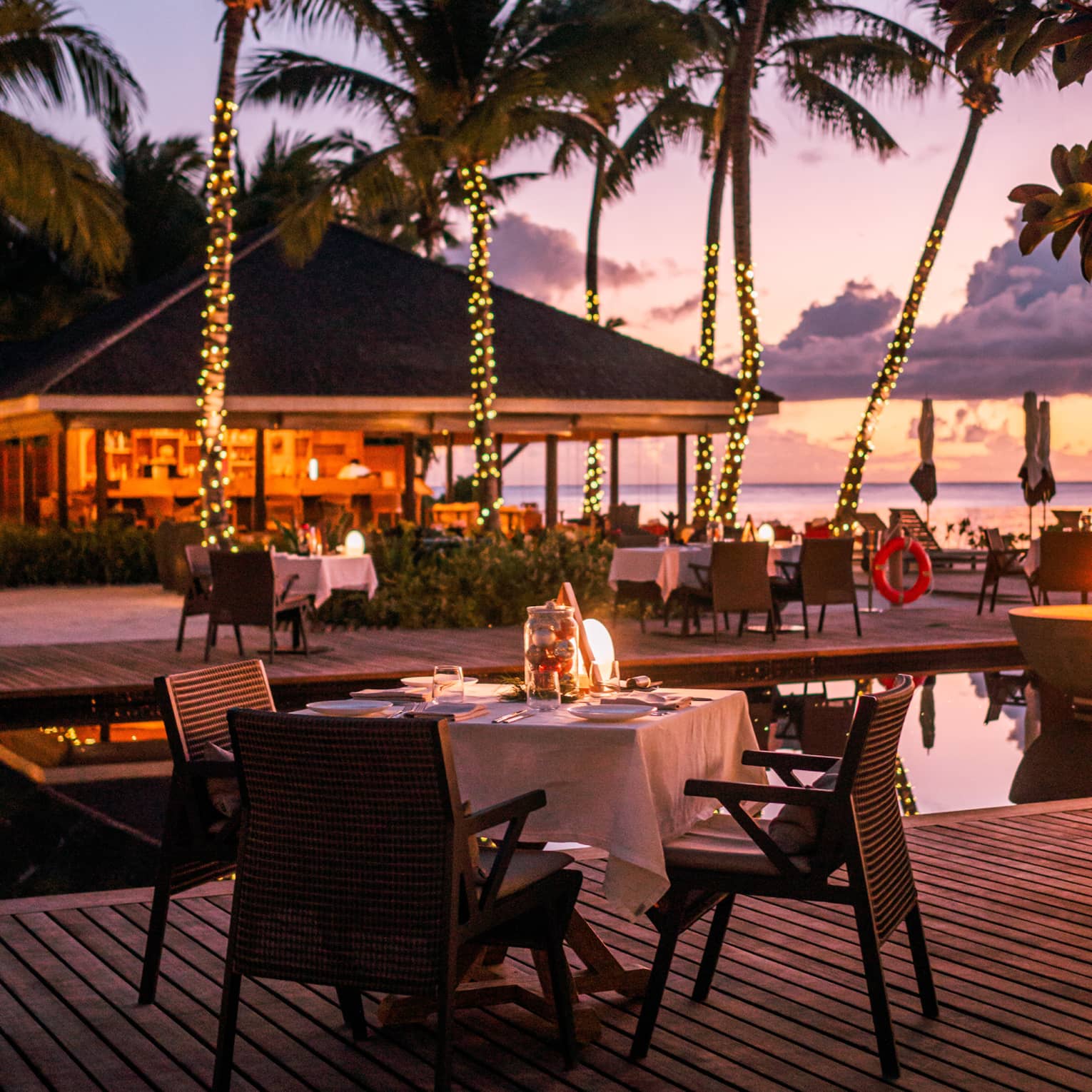 Claudine waterfront terrace with square dining tables amid palm trees wrapped in white lights at dusk