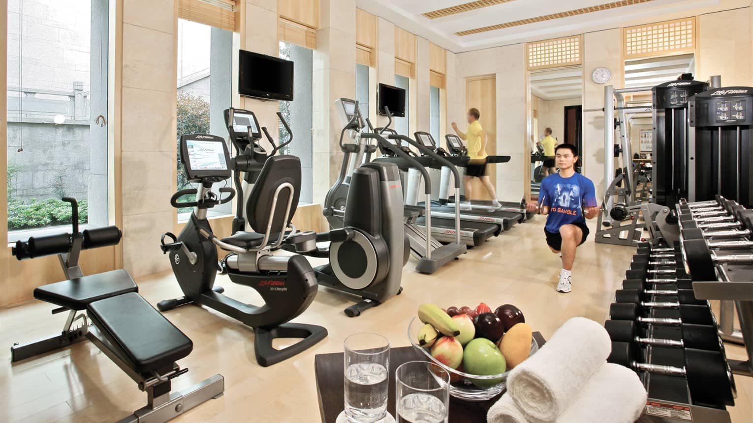 Man doing lunges in indoor gym, cardio machines line a wall of windows, free weights