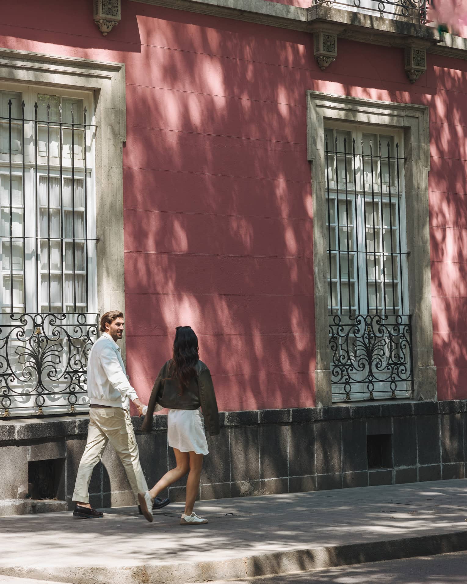 Couple strolls beside pastel pink bricked building featuring tall, paned windows adorned with ornate wrought iron railings.