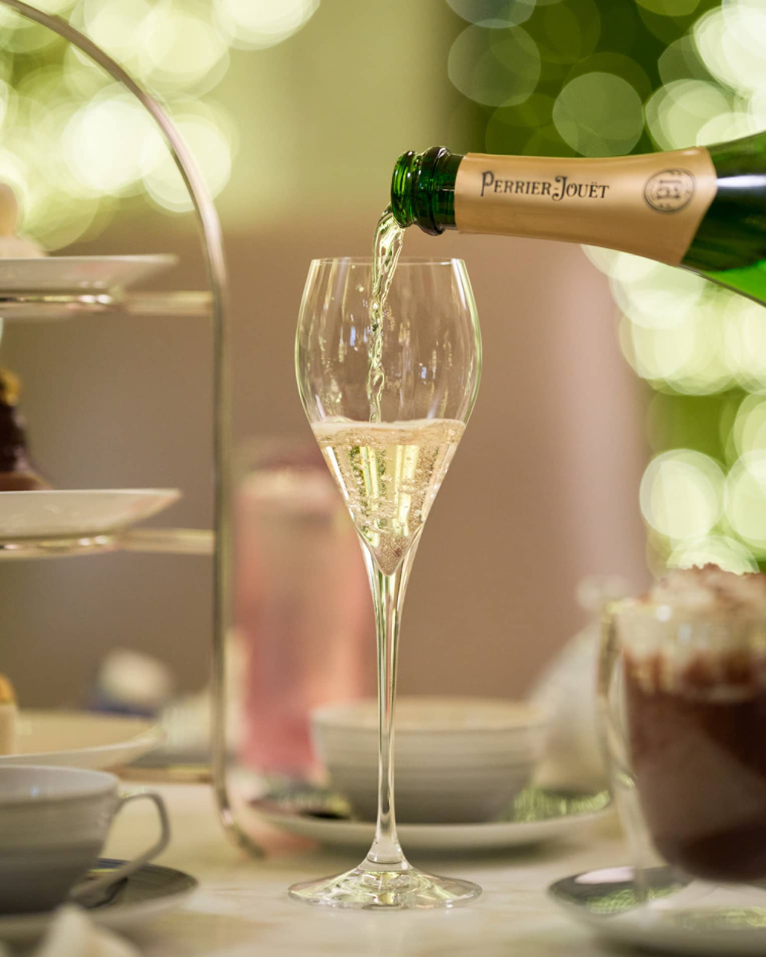 Champagne is poured from a bottle of Perrier Jouet into a glass next to a tiered tray of treats and a frothy cup of espresso.