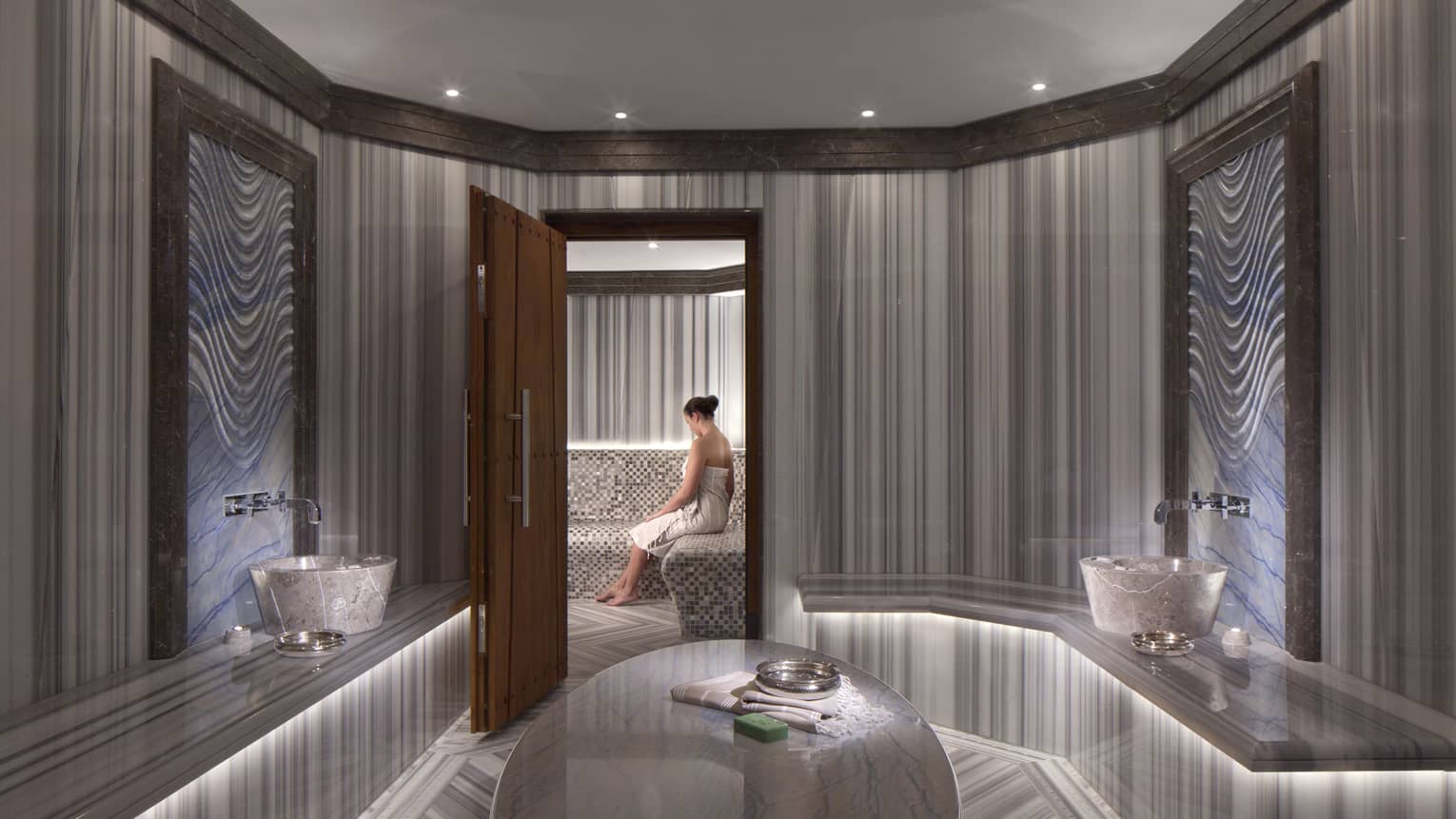Elegant marble spa room with open wood door where woman wearing towel sits on bench 