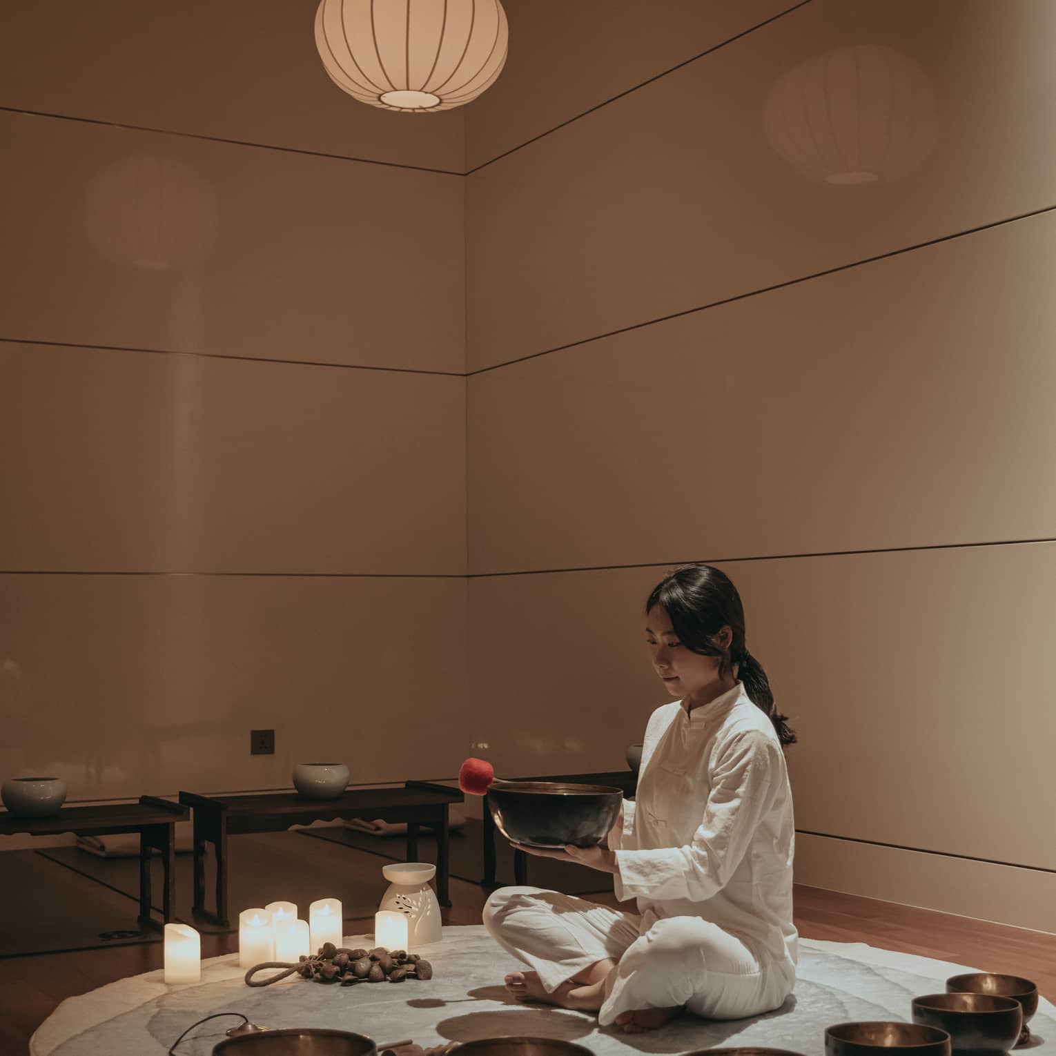 In a dim room, cross-legged on a circular rug amid various candles and bowls, a barefoot practitioner holds a bowl and mallet.
