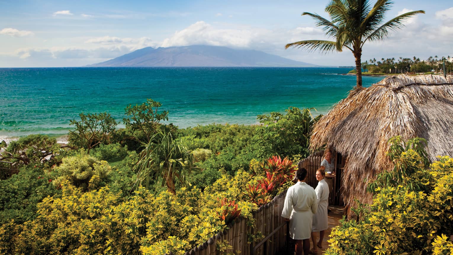 Couple in white bathrobes follow spa attendant into thatched-roof hut on path looking out at ocean