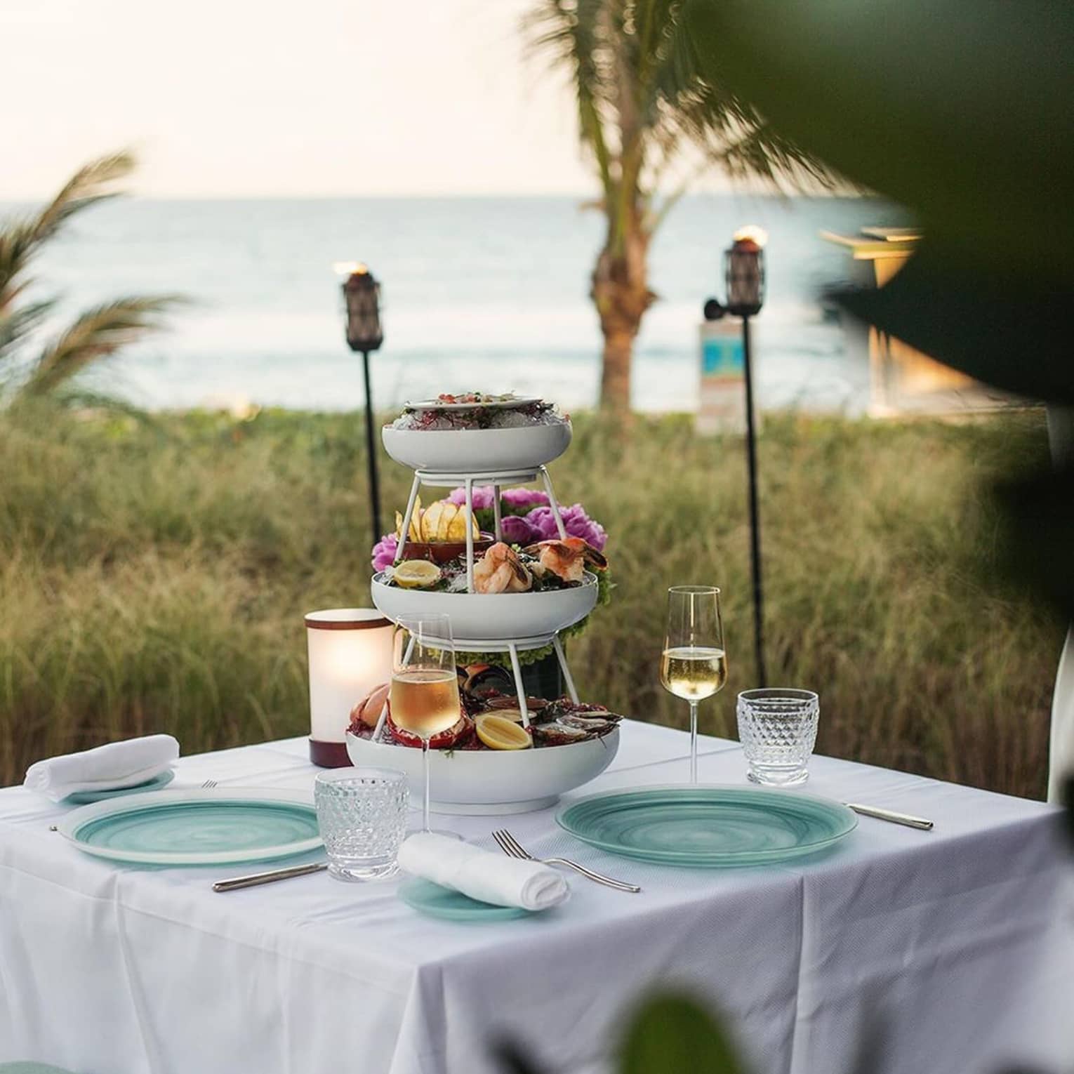 Three-tiered seafood tower is set in the centre of a dining table set outside with the beach nearby