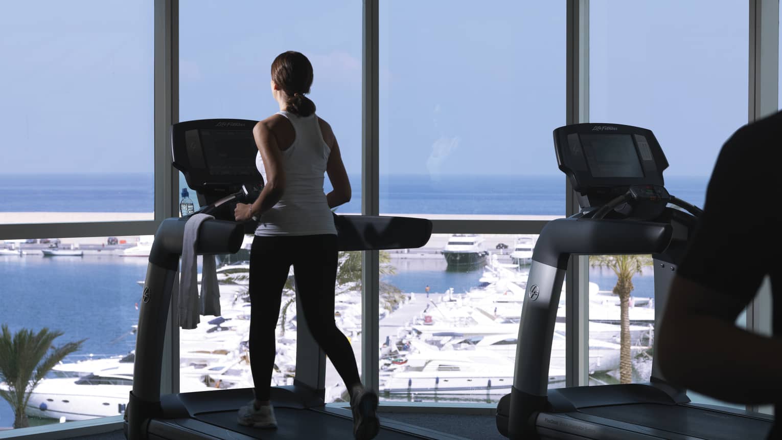 Back of woman in white sleeveless workout shirt and black pants runs on treadmill, faces window