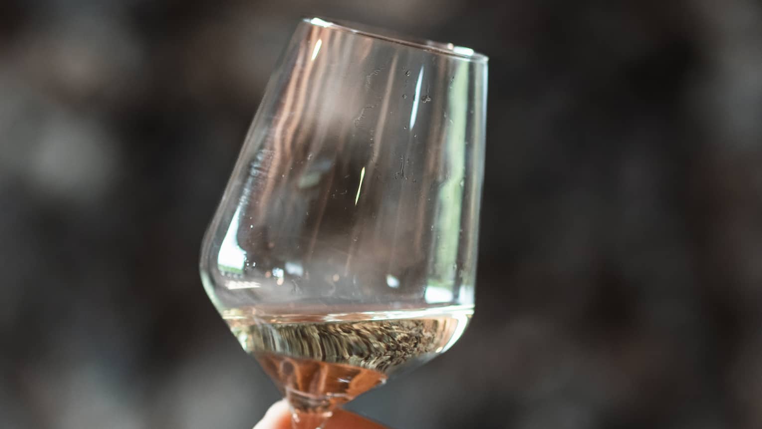A hand lightly grips the stem of a glass of white wine, swirling it on a slight angle.